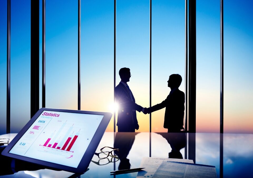 Silhouettes of two business professionals shaking hands in front of a large window with a sunset view, tablet displaying statistics and data analysis in the foreground, symbolizing B2B marketing and content syndication strategies.